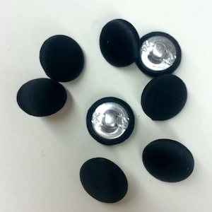 Könnte beinhalten: Acht schwarze, stoffbezogene Knöpfe mit silbernen Metallrücken. Die Knöpfe sind rund und haben eine glatte Oberfläche.