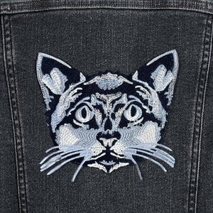 May include: A white embroidered cat face patch on a dark denim jacket.