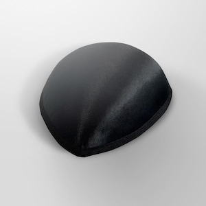 May include: A black satin sleep mask with a rounded shape and a black fabric trim. The mask is designed to block out light for improved sleep quality. The smooth, shiny texture of the satin is visible.