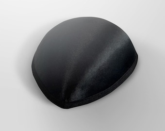May include: A black satin sleep mask with a rounded shape and a black fabric trim. The mask is designed to block out light for improved sleep quality. The smooth, shiny texture of the satin is visible.