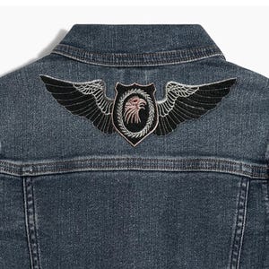 May include: A dark blue denim jacket with an embroidered patch on the back. The patch features a black eagle with wings spread, inside a circular frame with a shield shape.
