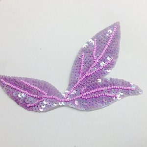 May include: A pair of purple iridescent sequins in the shape of leaves. The sequins are sewn together with pink thread.