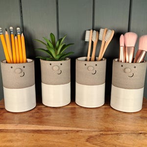 May include: Four ceramic containers with smiling faces, each with a grey top and white bottom. One holds pencils, another a small plant, the third toothbrushes, and the last makeup brushes. The containers are on a wooden surface.
