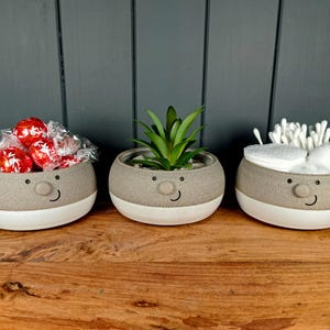 May include: Three small, round ceramic pots with smiling faces. One contains wrapped red sweets, another a small succulent plant, and the third cotton buds and pads. The pots are grey with white bases, set on a wooden surface.