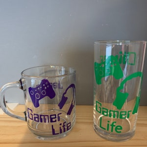May include: Two clear glass cups with the words "Gamer Life" printed on them. One cup is a mug with a purple video game controller and headphones, the other is a taller glass with a green video game controller and headphones.