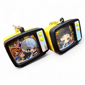 May include: Two yellow and black miniature television keychains. Each keychain features a cartoon image on the screen. The televisions have yellow buttons and a white side panel. The keychains have a silver metal ring.