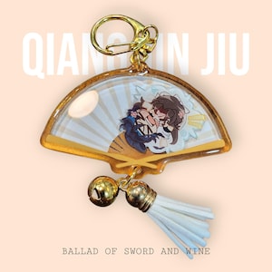 May include: A fan-shaped keychain with a gold-colored frame and a cartoon illustration of two characters. The keychain has a gold-colored clasp, a gold bell, and a white tassel. The text "QIAN QIN JIU" and "BALLAD OF SWORD AND WINE" are visible.