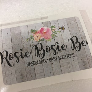May include: A white and grey woodgrain background with a floral design and a bee. The text "Rosie Bossie Be" is in black cursive font. Below the text is the phrase "Handmades - Baby Boutique".