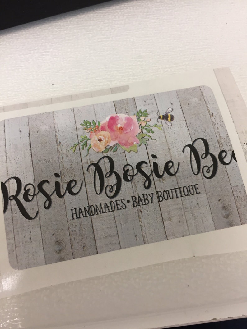 May include: A white and grey woodgrain background with a floral design and a bee. The text "Rosie Bossie Be" is in black cursive font. Below the text is the phrase "Handmades - Baby Boutique".