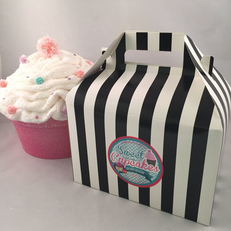 May include: A black and white striped cardboard box with a handle. The box has a sticker that says "Sweet Cupcakes" with a cupcake graphic. The box is next to a large white cupcake with pink frosting and colorful sprinkles.