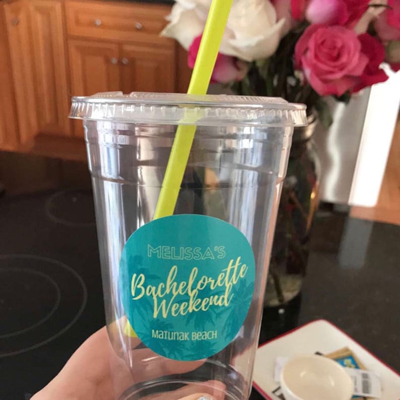 Puede incluir: Un vaso de pl&aacute;stico transparente con una pajita amarilla y una pegatina azul verdoso y dorada que dice "Melissa's Bachelorette Weekend Matunuk Beach".