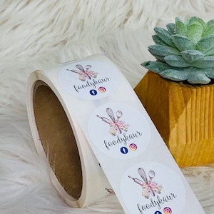 Op de afbeelding: Rol witte ronde stickers met een logo-ontwerp met een deegroller, garde en lepels met roze bloemmotieven. De logo-tekst is "foodykaur" met Facebook- en Instagram-iconen.
