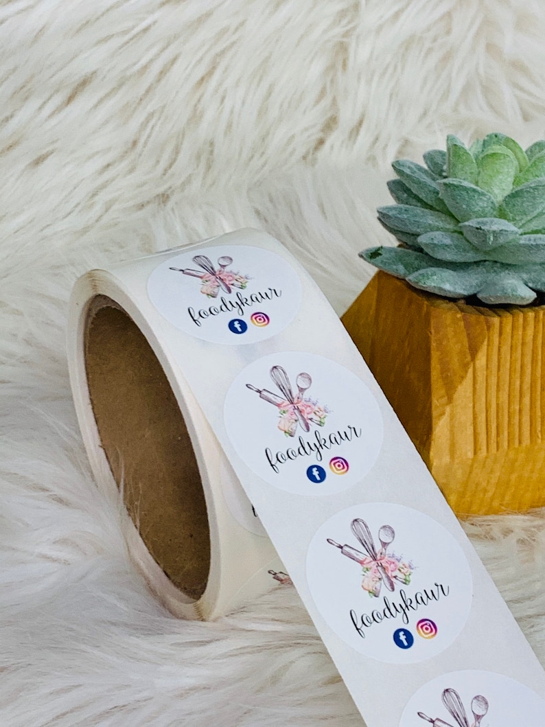 Op de afbeelding: Rol witte ronde stickers met een logo-ontwerp met een deegroller, garde en lepels met roze bloemmotieven. De logo-tekst is "foodykaur" met Facebook- en Instagram-iconen.