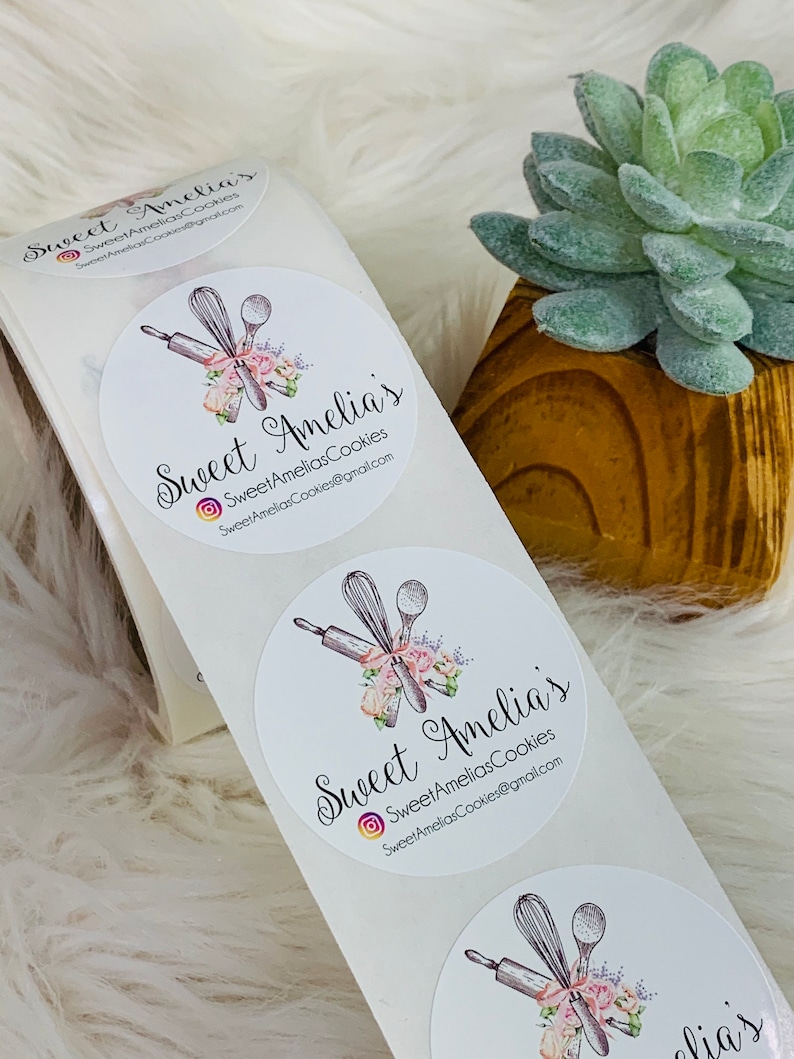 Op de afbeelding: Drie witte ronde stickers met een bloemmotief en de tekst "Sweet Amelia's" en "SweetAmeliasCookies@gmail.com".