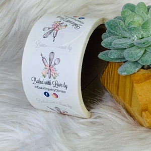 Op de afbeelding: Een rol witte ronde stickers met een bloemmotief en de tekst "Baked with Love by @CakePopsbyDivine" en social media-iconen voor Facebook en Instagram.