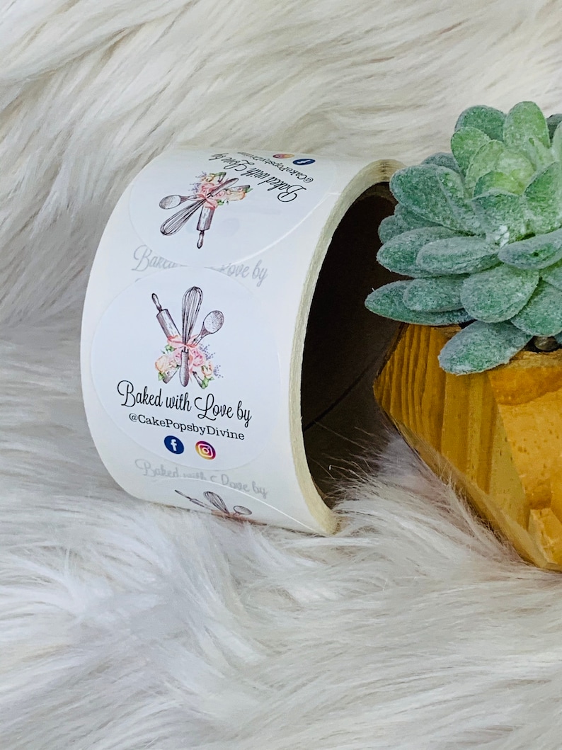 Op de afbeelding: Een rol witte ronde stickers met een bloemmotief en de tekst "Baked with Love by @CakePopsbyDivine" en social media-iconen voor Facebook en Instagram.
