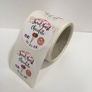 May include: Roll of round stickers with a pink and white design. The stickers feature the text "Sweet Heart Chocolate" with a pink heart pattern and a lollipop graphic. The phone number 917-561-9810 is also included on the sticker.