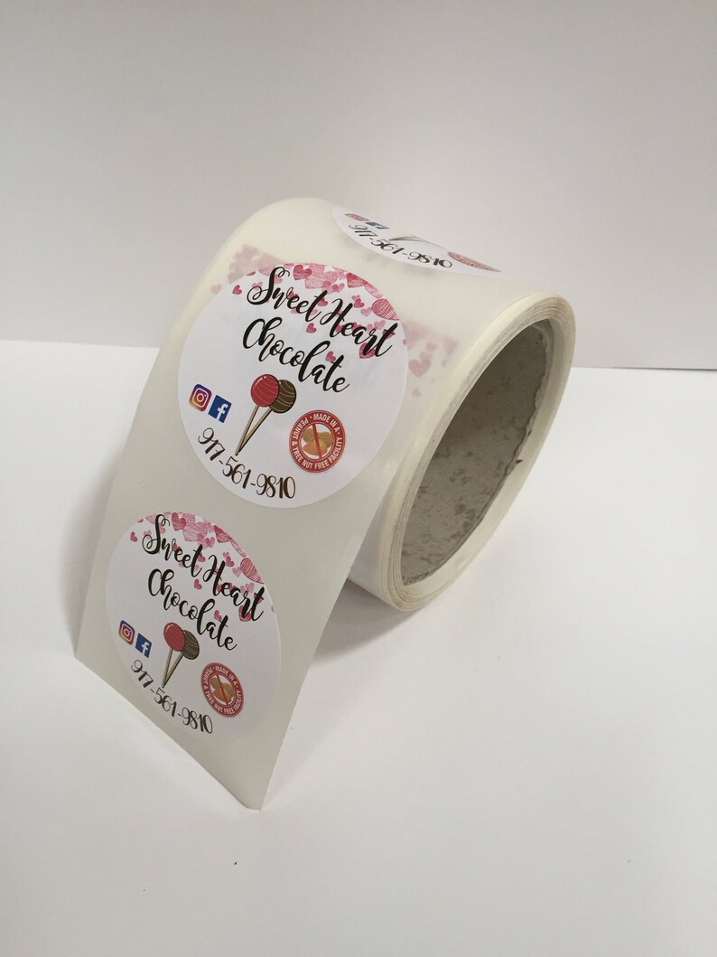 May include: Roll of round stickers with a pink and white design. The stickers feature the text "Sweet Heart Chocolate" with a pink heart pattern and a lollipop graphic. The phone number 917-561-9810 is also included on the sticker.
