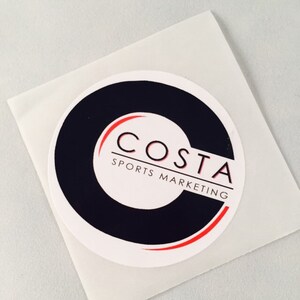 Puede incluir: Una pegatina circular blanca con un logotipo negro y rojo. El logotipo es una "C" estilizada con la palabra "COSTA" en letras rojas y las palabras "SPORTS MARKETING" en letras negras debajo.