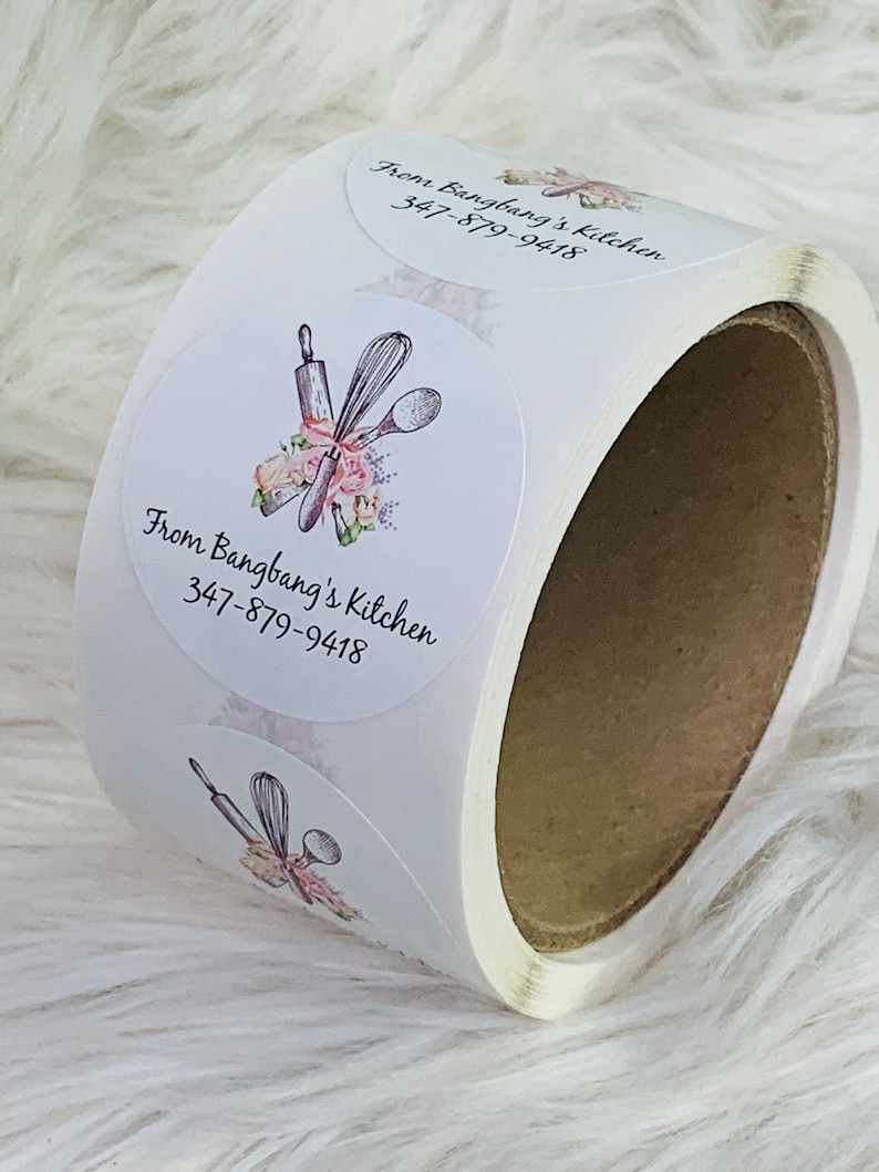 Op de afbeelding: Rol witte ronde stickers met een roze bloemenontwerp en keukengerei. De tekst op de sticker luidt "From Bangbang's Kitchen 347-879-9418".