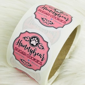 Puede incluir: Rollo de pegatinas redondas rosas y blancas con el texto "Hunybear Sugar Cookies" y un logotipo de huella de pata.