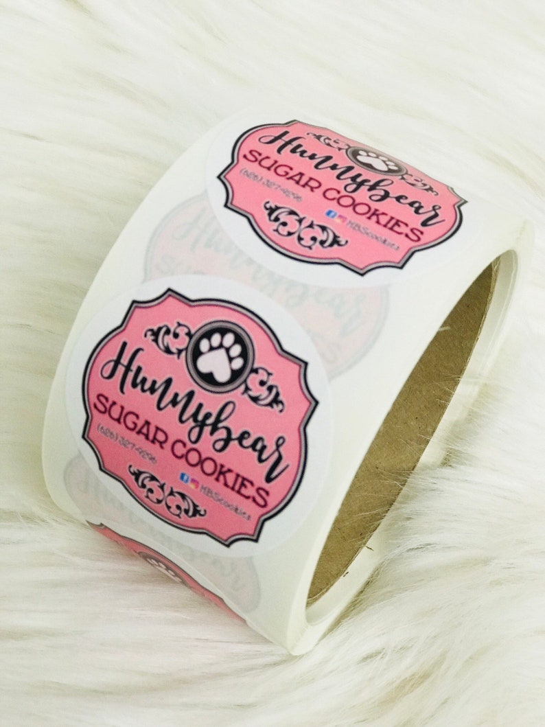 Puede incluir: Rollo de pegatinas redondas rosas y blancas con el texto "Hunybear Sugar Cookies" y un logotipo de huella de pata.