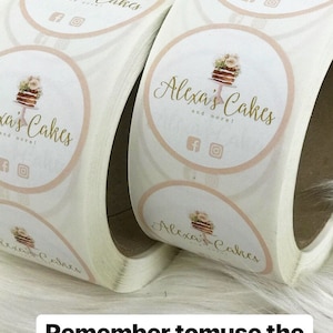 Puede incluir: Un rollo de pegatinas redondas con fondo blanco y un c&iacute;rculo rosa en el centro. El c&iacute;rculo presenta una ilustraci&oacute;n de acuarela de un pastel con flores y el texto "Alexa's Cakes and more!" en escritura dorada. El texto "Remember to use the codes from our influencers to help them" est&aacute; escrito en negro debajo del rollo de pegatinas.