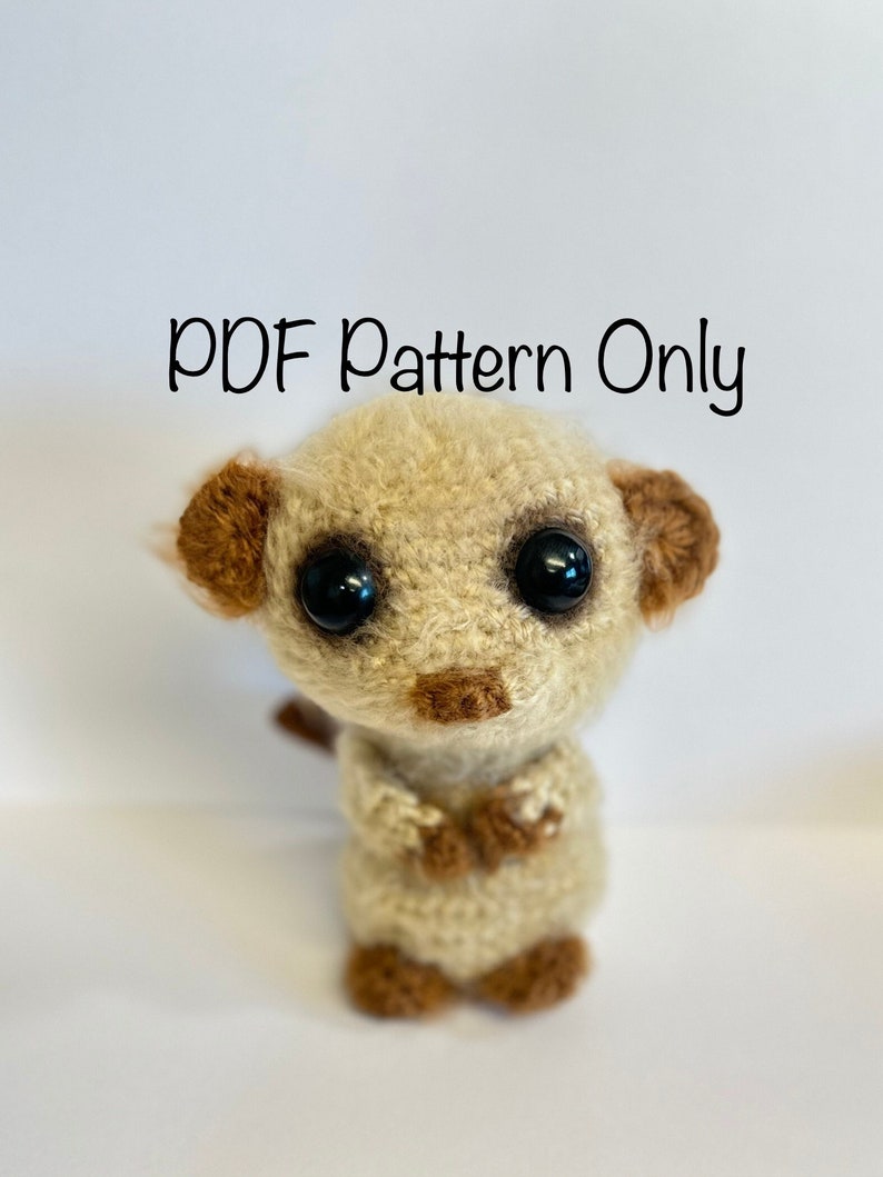 May include: A crocheted amigurumi meerkat toy with brown ears, black eyes, and a brown nose. The toy is standing on a white background. The text "PDF Pattern Only" is written above the toy.