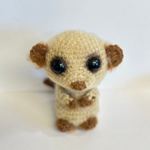 May include: A crocheted meerkat stuffed animal with brown ears and black eyes. The meerkat is sitting upright with its paws crossed in front of it.