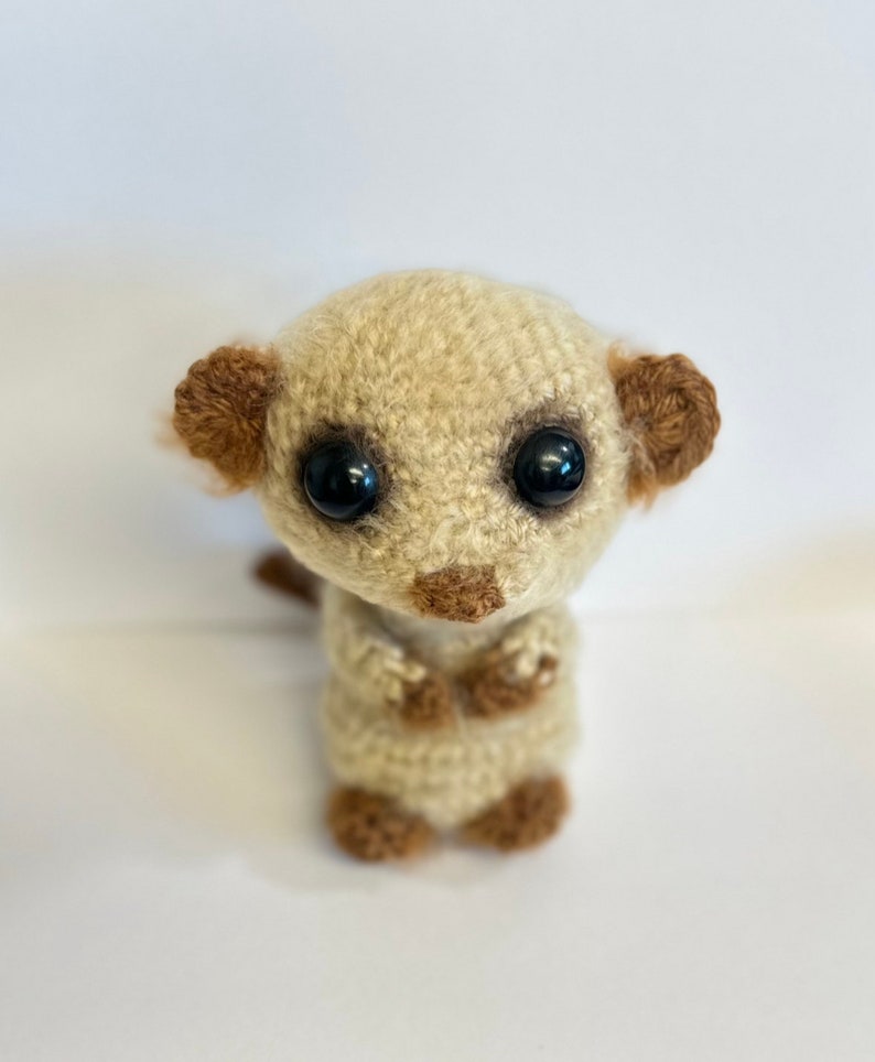 May include: A crocheted meerkat stuffed animal with brown ears and black eyes. The meerkat is sitting upright with its paws crossed in front of it.