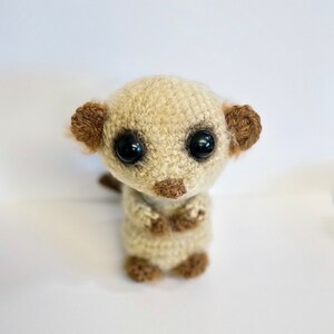 May include: A crocheted meerkat toy with brown ears and black eyes. The toy is standing upright with its arms crossed.