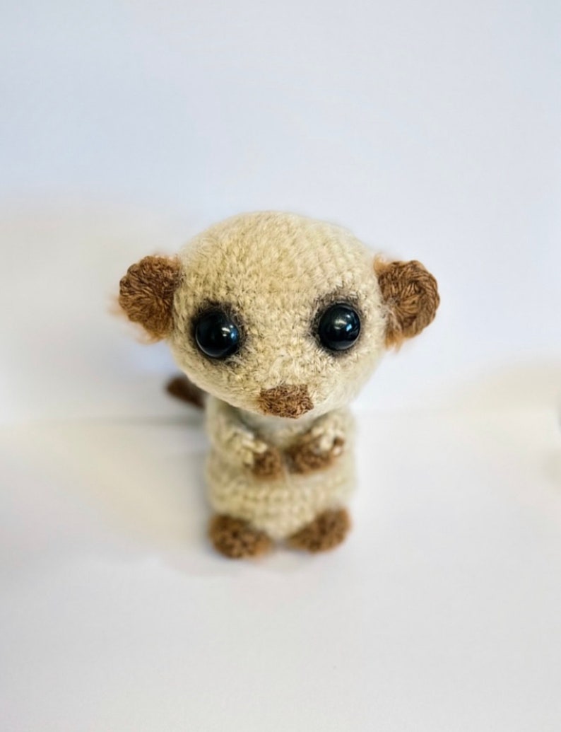 May include: A crocheted meerkat toy with brown ears and black eyes. The toy is standing upright with its arms crossed.