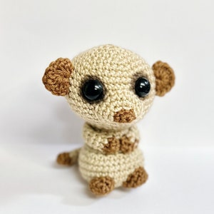May include: A small crocheted meerkat toy with brown ears, black eyes, and a beige body. The meerkat is sitting upright with its arms crossed.