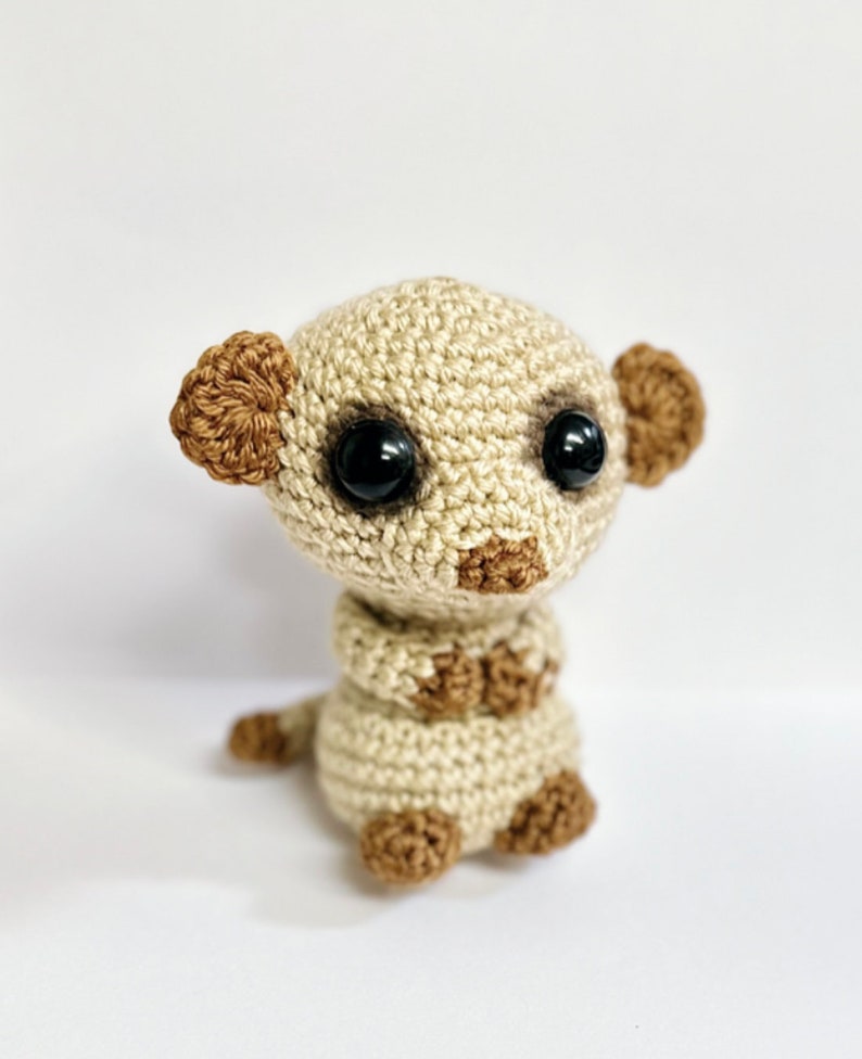 May include: A small crocheted meerkat toy with brown ears, black eyes, and a beige body. The meerkat is sitting upright with its arms crossed.