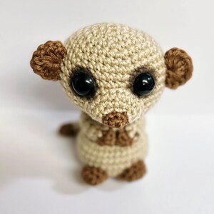 May include: A crocheted meerkat toy with brown ears and black eyes. The meerkat is sitting upright with its paws tucked in.