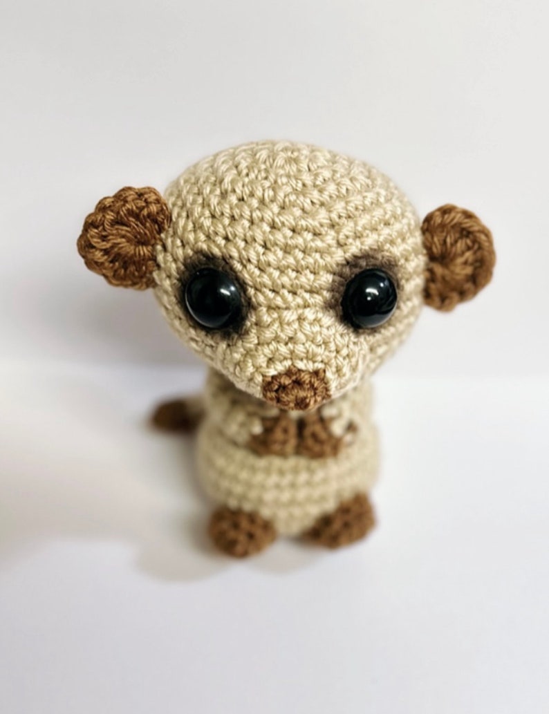 May include: A crocheted meerkat toy with brown ears and black eyes. The meerkat is sitting upright with its paws tucked in.