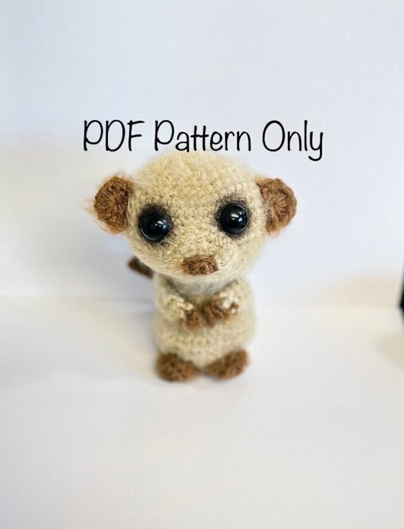 May include: A crocheted amigurumi meerkat toy with brown ears and black eyes. The toy is standing on a white surface. The text "PDF Pattern Only" is written above the toy.