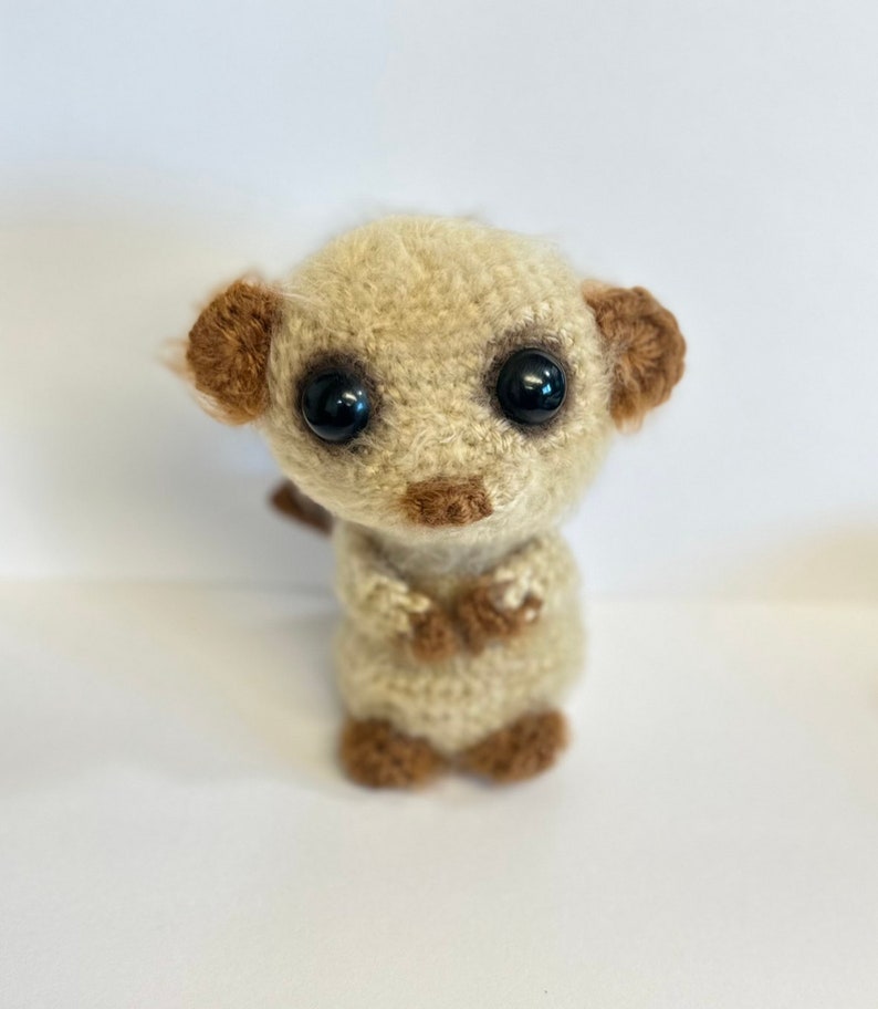 May include: A crocheted stuffed animal resembling a meerkat with brown ears and black eyes. The meerkat is standing on a white surface.