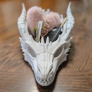 May include: A white dragon head-shaped container holding keys, a car key fob, and a pink heart-shaped item. The container has detailed scales, horns, and eye sockets. The container is sitting on a wooden surface.