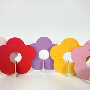 May include: Five colorful flower-shaped decorations in shades of beige, red, purple, yellow, and pink. Each flower has a white center and is mounted on a wooden stick. The flowers are arranged against a white background.