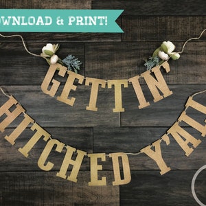 Puede incluir: Una pancarta rústica de arpillera marrón con el texto "Gettin' Hitched Y'all" enhebrada en una cuerda. La pancarta está decorada con flores blancas y vegetación.