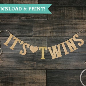 May include: A brown burlap banner with the text "It's" followed by a heart and then "Twins". The banner is strung with twine and is hanging against a wood background.