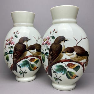 May include: Two white ceramic vases. Each vase features a painted design of brown birds on a branch with pink flowers and green leaves. The vases have a bulbous body and a flared top.