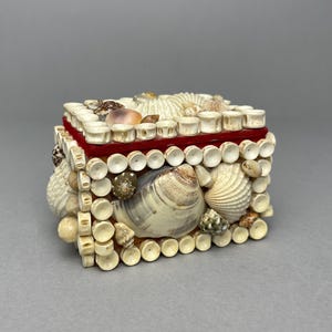 May include: A decorative box, rectangular in shape, covered in seashells. The box has a red velvet lining and is embellished with small, cylindrical pieces and a variety of shells in white, beige, and brown hues.
