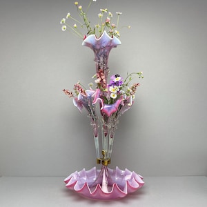 May include: A multi-tiered, pink glass vase with a ruffled design, filled with fresh flowers. The vase features a central column with multiple flared openings and a wide, scalloped base. The flowers include daisies, purple, and pink blooms, creating a delicate and elegant display.
