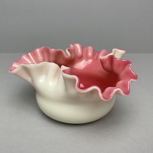 Könnte beinhalten: Eine kleine, gerüschte Schale mit weißem Boden und rosa Innenraum und Rand. Der Rand hat einen wellenförmigen, gewellten Rand. Die Schale ist wahrscheinlich aus Keramik oder Porzellan und steht vor einem neutralen Hintergrund.