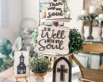 May include: A three-tiered tray decorated with a variety of wooden signs featuring religious quotes. The signs include the phrases "It is Well With My Soul", "Then Sings My Soul", and a cross. The tray is decorated with greenery and a small church-shaped sign.