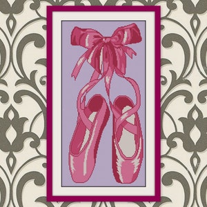 May include: Cross-stitch artwork of pink ballet pointe shoes with ribbons tied in a bow. The shoes are set against a light purple background, framed in a magenta border. The artwork is displayed against a patterned wallpaper.
