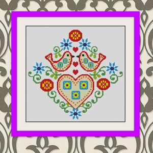 Könnte beinhalten: Ein gerahmtes Kreuzstichdesign mit zwei roten Vögeln, die sich über einem Herzen gegenüberstehen. Das Herz und die Vögel sind von blauen Blumen, grünen Ranken und roten und gelben kreisförmigen Akzenten umgeben. Das Kunstwerk ist in einem weißen Passepartout und einem lila Rahmen gerahmt.