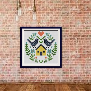 May include: A framed cross-stitch featuring a folk art design. Two black birds flank a yellow house, with a red heart above. The design is surrounded by green leaves and blue flowers, set against a white background.